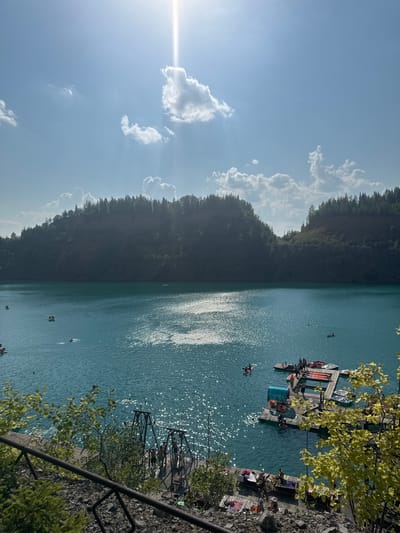 The image depicts a scenic view of Karyer Izvestnyak, Russia, a former limestone quarry now transformed into a popular recreational area. The water is a striking turquoise color, reflecting the bright sunlight that beams down from a clear blue sky. Scattered clouds add depth to the sky, with the sun's rays piercing through in a dramatic effect.
The lake is dotted with various watercraft, including paddleboats and inflatable rings, indicating a day of leisure activities. Several people are visible enjoying the water, swimming or relaxing on small boats. A wooden dock extends into the lake, equipped with multiple paddleboats and some individuals socializing on the platform. Another smaller dock to the left has a swing-like apparatus and a few people gathered.
The surrounding landscape is characterized by steep, tree-covered hillsides, characteristic of a quarry environment. Lush green foliage frames the foreground, suggesting a vibrant natural setting. The overall impression is one of a tranquil and inviting summer day, with the clear water and sunny skies creating an idyllic atmosphere.
