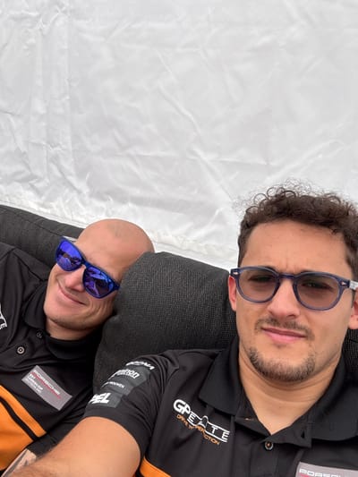 The image shows two men sitting side-by-side on a black couch. The man on the left is wearing a black polo shirt with an orange stripe and a white logo that reads "Porsche Team GP DEUTSCHLAND". He is wearing blue sunglasses and is smiling. He is leaning back with his head resting on the couch. The man on the right is wearing a black polo shirt with an orange stripe and a white logo that reads "G-P-E-A-T-E DRIVE TO PERFECTION". He is wearing blue glasses and has a serious expression. The image is taken in Malmedy, Belgium, at the coordinates 50.442, 5.971. The environment is a bright, white tent with a slightly wrinkled, white fabric background. The image is likely a selfie.