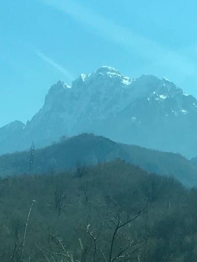 The image displays a mountainous landscape observed from Donja Jablanica, Bosnia and Herzegovina. The scene is dominated by a multi-layered vista under a light blue sky exhibiting faint, wispy cloud formations. In the furthest background, a large, rugged mountain range features distinct, snow-capped peaks. These distant mountains appear somewhat obscured by atmospheric haze. In the midground, a series of lower hills covered with deciduous trees, largely bare of foliage, are present. A tall, slender lattice structure, resembling a utility pylon or communications tower, is discernible on the slope of one of these midground hills, positioned towards the left. The immediate foreground consists of bare branches from closer deciduous trees, partially obscuring the base of the midground hills. No individuals, animals, or specific actions are depicted within the frame.