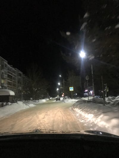 A dark street scene, likely at night, shows a road covered in a slushy, icy mix, with piled snow to the side. A streetlamp illuminates the road. A building stands to the left. Various traffic signs are visible along the road, including a crosswalk sign. A vehicle appears to be parked on the right side of the street. The image is taken from inside a car, with part of the car’s interior visible on the right.
