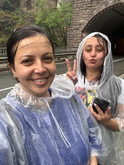 This vertical selfie captures two young women outdoors, dressed in clear plastic rain ponchos, suggesting a rainy day. The image appears to be taken at the Bachkovo Monastery in Bulgaria, indicated by the location context and the rustic stone archway visible behind them.
The woman in the foreground, positioned at the bottom right of the frame (when viewed upright), has dark, wet hair and is smiling warmly, looking directly at the camera. She wears a blue garment under her poncho, which is speckled with raindrops.
Behind her, towards the top left, the second woman also has dark, wet hair and is making a playful "duck face" while holding a smartphone in a clear case with her left hand. Her right hand displays a peace sign. She wears a white t-shirt with a colorful graphic design beneath her rain gear, also covered in water droplets.
The background features a distinctive stone archway or tunnel entrance on the left, with a red and white striped barrier partially visible inside. Lush green trees and natural rock formations are discernible further back, indicating a scenic, possibly mountainous or historical, outdoor setting. A metal railing or fence is also partially visible, suggesting a walkway or bridge. Despite the rain, both women convey a cheerful and lighthearted mood. No discernible text is clearly visible.