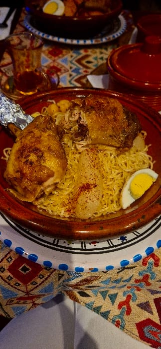 A plate of Moroccan chicken with seffa. The golden-brown chicken pieces sit atop a bed of fine vermicelli noodles, garnished with a half-boiled egg and a sprinkling of spice. The plate is a traditional tagine dish with a patterned rim. Adjacent to the main dish, there's a smaller plate with another halved egg, a glass of tea, and a red teapot. The surrounding surfaces are covered in a colorful, geometric pattern.
