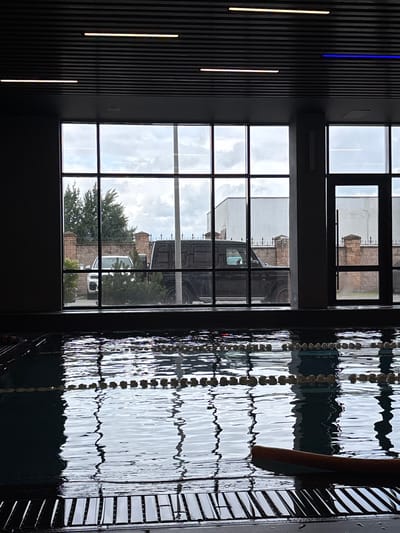 This is a photograph taken indoors, overlooking a swimming pool at the Monolit, Russia fitness club. The pool area features a lane divider made of buoys and ropes. In the foreground, a dark brown kickboard floats on the surface of the water. Beyond the pool, large windows offer a view of an outdoor area with two parked cars, one white and one black, and a brick fence. The ceiling of the indoor space is dark and features fluorescent lighting.