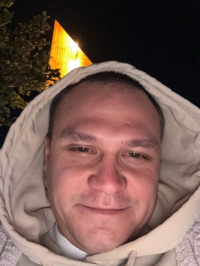 A close-up selfie shows a middle-aged man with short hair and light skin, wearing a light-colored hooded sweatshirt. His face is centered in the frame, slightly angled. His expression is relaxed. In the lower-left corner, a building is lit up by bright yellow light, partially obscured by greenery. The background is dark, suggesting night. It is not possible to verify this photograph's location in the city of Sindieti, Georgia.