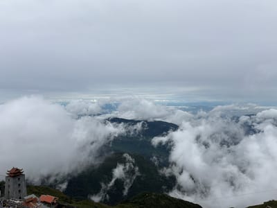 This image captures a breathtaking, cloudy mountain landscape. The primary subjects are the majestic mountains, shrouded in thick, white clouds, and a traditional stone tower with a red-tiled roof, situated on a lush, green mountainside. The tower appears to be a historical or cultural structure.

The scene depicts a serene, possibly cool, and misty atmosphere, suggesting a location at a high altitude. The sky is overcast, filled with layers of clouds that obscure the sun, creating a diffused, soft light. The mountains are covered in dense, dark green foliage, hinting at a tropical or subtropical mountainous region. In the distance, the clouds part slightly, revealing glimpses of more distant mountain ranges and possibly a valley with scattered settlements.

Notable details include the presence of what appears to be a cable car system, with cables stretching across the frame and small cabins visible, indicating that this is a popular tourist destination or a location accessible by cable car. The presence of a few people near the base of the tower suggests it's an accessible point of interest.

The overall mood conveyed is one of awe and tranquility, with the vastness of the cloudy mountain range evoking a sense of wonder. The time of day is indeterminate due to the cloud cover, but the lighting suggests it could be mid-morning or afternoon. The location cues, such as the architecture of the tower and the lush vegetation, combined with the context of "Ban Bo, Vietnam," strongly suggest this is Fansipan mountain in Vietnam. No visible text is present in the image.