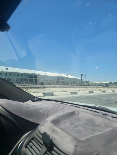 This is a photograph taken from inside a vehicle, looking out through the windshield at a light blue, multi-story building. The building appears to be commercial or institutional, with many windows and a long, sloping roof. There is a water tower to the right of the building, and a fence surrounds the property. The landscape is dry and sandy, with sparse vegetation. The sky is clear and blue. The photograph appears to be taken during the day. The location is stated as Taylakan, Uzbekistan.
