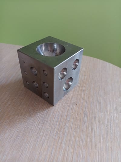 A die-shaped metal object with several circular indentations on its faces sits on a light-colored wooden table. The object has a large concave circular indentation on its top surface. The background is a solid light green color. The image does not provide any information about the city of Vilyuysk, Russia.