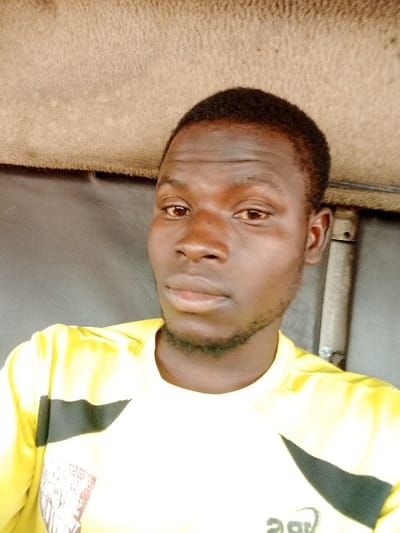 A close-up, chest-up portrait captures a young adult male in Kadawa, Nigeria. The individual has dark skin, short dark hair, and a neatly trimmed dark beard. He is wearing a bright yellow, short-sleeved, crew-neck t-shirt featuring black and white graphic designs on the shoulders/chest. His expression is neutral as he looks directly into the camera. The background consists of a light brown, textured surface in the upper portion, likely a ceiling or interior lining, and a darker, plain surface below it. A metallic vertical structure is partially visible on the right side of the frame.