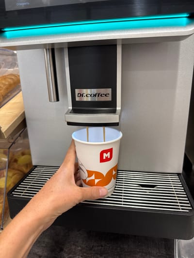 A person's hand is holding a paper coffee cup under a dispenser on a Dr. Coffee machine. The cup has an orange and white pattern and a red 'M' logo. The machine has a silver and black finish, with a bright blue light strip on the left side. The dispenser area has a metal grate. The background shows a display case with baked goods. The image is likely taken indoors, possibly in a cafe or convenience store. There is text on the machine that says "dr.coffee" and on the blue light strip which says "Отменить" (Cancel in Russian). The setting appears to be Kuivozi, Russia. The activity is the dispensing of coffee into a cup.
