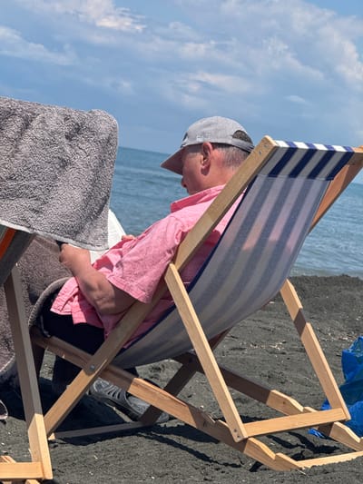 A man wearing a grey baseball cap and a pink collared shirt is sitting in a striped deck chair on a dark sand beach in Shekvetili, Georgia. He is holding a book or magazine and appears to be reading. A grey towel is draped over the arm of the deck chair. The sea is visible in the background under a partly cloudy sky.