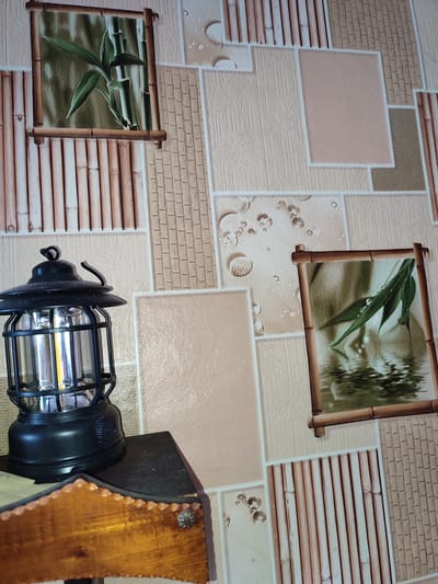 The image depicts an indoor scene with a wallpapered wall and a lantern on a piece of furniture. The wallpaper features a mosaic of textured panels, some with bamboo motifs, others with a light beige textured pattern, and some with images of bamboo leaves and water. Two framed pictures of bamboo leaves are visible on the wall. To the left, a black lantern with a metal cage and a bright LED light is placed on a dark surface, which is part of a wooden object with a textured brown finish. The setting is a room in a house or apartment in Voskobiinyky, Ukraine. There are no people visible in the image.