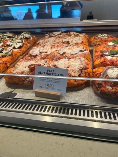 The image displays a close-up of various Italian pizzas, specifically "pizza alla pala" (pizza by the shovel), showcased in a refrigerated or heated display case within what appears to be a food establishment in Dorno, Italy.
The main subject is a large rectangular pizza labeled "PALA ALL'AMATRICIANA." This pizza features a thick, golden crust and is generously topped with a red sauce, chunks of what is identified as guanciale (cured pork cheek), and a generous dusting of grated Pecorino Romano DOP cheese and black pepper.
Above the Amatriciana pizza, there are other types of pizza. One visible portion features anchovies and fresh green basil leaves on a red sauce base, while another has anchovies on a creamy white base, possibly with a dollop of soft cheese. Below the Amatriciana pizza, a section of another pizza is visible, topped with green herbs and white grated cheese.
A white price tag with a wooden base stands in front of the "PALA ALL'AMATRICIANA." The visible text on the tag reads:
- "PALA ALL'AMATRICIANA"
- "Pizza"
- "pecorino romano DOP e pepe nero"
- "Pizza alla pala with tomato pulp, guanciale, PDO pecorino romano and black pepper"
- "€ 8,20"
Below the price, there is smaller, illegible text, likely detailing ingredients or allergy information. The overall scene suggests a deli or bakery counter where fresh pizzas are prepared and sold.