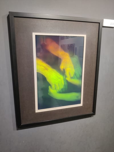 The image shows a framed artwork hanging on a grey wall in Thessaloniki, Greece. The artwork is a photograph with a prominent display of hands, rendered in vibrant yellow and green hues, creating an abstract and somewhat ethereal effect. Two sets of hands are intertwined, with one pair appearing to be helping or holding the other. The background of the photograph is dark and blurred, with some faint reflections of what appear to be circular shapes. A small white label is attached to the wall to the right of the frame, with Greek text and the English translation "HANDS OF HOPE." The lighting in the scene is ambient, with some light reflecting off the glass of the frame. The overall impression is one of a contemporary art exhibition.