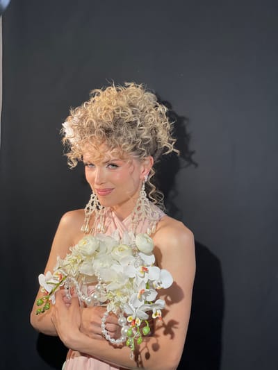 A woman with curly blonde hair and a pale pink dress holds a bouquet of white flowers and pearls, standing in front of a black backdrop. She is smiling and looking down at the flowers. The scene is a portrait studio setting, possibly located in or referencing a location with significance to Vysokaya Gora, Russia, considering the request.
