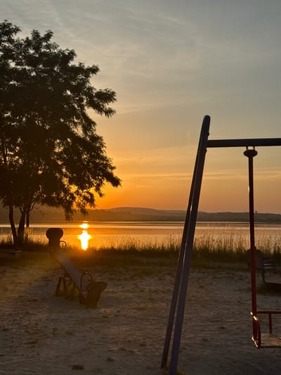 The image depicts a serene outdoor scene at sunset, likely a playground near a body of water. The sun, a brilliant orb of light, is low on the horizon, casting a warm golden glow across the sky and reflecting intensely on the water's surface. Tall reeds or marsh grasses line the water's edge, creating a natural border between the water and the sandy or gravelly ground.

In the foreground, the silhouette of a tree with dense foliage is visible, its branches reaching towards the sky. To the right, parts of a playground structure can be seen, including a seesaw. The metal frame of a swing set or climbing frame is also present, partially obscuring the view of the sunset. The ground appears to be sandy or composed of fine gravel, typical of a beach or park area.

The overall mood is peaceful and tranquil, evoking a sense of calm associated with the end of the day. The soft lighting and muted colors suggest a pleasant, possibly warm, weather condition. The time of day is clearly sunset, characterized by the low sun and the warm, saturated hues in the sky. The presence of playground equipment indicates a public recreational space, likely a park or a designated play area.

No visible text is present in the image. The location context provided is Berezhany, Ukraine, which aligns with a natural landscape that could feature a lake or river and recreational facilities. The image captures a moment of natural beauty enhanced by the presence of human-made structures.