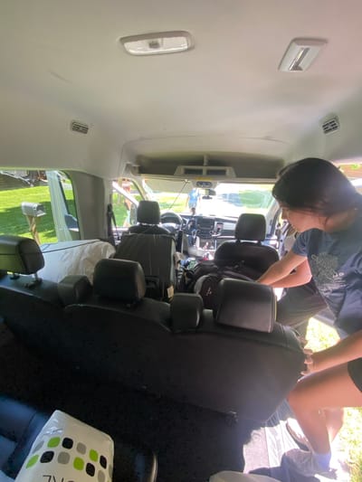 In Washington Township, a group of individuals are packing a van. The scene is set outdoors, with lush green grass and trees visible in the background, suggesting a suburban or rural location. The van's interior is packed with various items, including what appears to be bedding and luggage. One woman, wearing a blue t-shirt, is actively involved in the packing process, reaching into the van. Another person is visible in the driver's seat, and a third individual stands near the open rear door. The overall impression is one of preparation for a trip or move, with a focus on efficient organization of belongings within the vehicle.