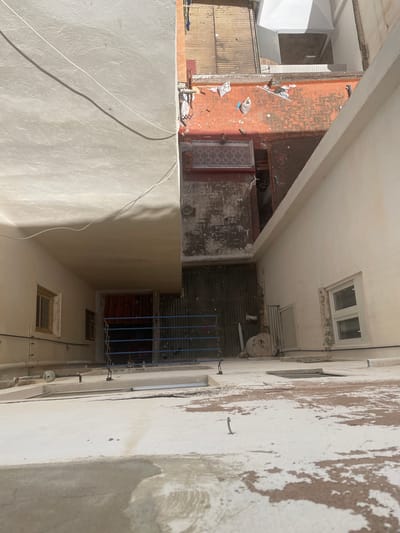 The image depicts an outdoor courtyard or alleyway, likely in Sagunto, Spain, given the location context. The scene is cluttered with various objects, suggesting a disused or storage area. There are several structures visible: a building with a weathered, peeling facade on the right, and another building with an orange, brick-like exterior and wooden siding on the left. A cream-colored, textured wall dominates the foreground, with a white electrical wire casting a shadow across it.
In the background, the courtyard reveals more details. There's a corrugated metal roof or wall, and a blue metal grate or fence. Various items are scattered around, including bags, pipes, and what appear to be building materials or debris. Small windows with metal grates are visible on the buildings, and a small, white air conditioning unit or fan is mounted on the wall. The lighting suggests it is daytime, with harsh sunlight casting strong shadows. The overall impression is one of an unkempt, possibly abandoned or neglected urban space. There is no visible text in the image. The scene does not appear to involve people or any specific event.