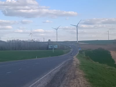 The image presents a wide view of a rural landscape featuring a winding asphalt road, agricultural fields, and numerous wind turbines under a partly cloudy sky. The two-lane road, with white dashed markings and a solid white line on the shoulder, extends from the bottom of the frame and curves to the right before disappearing into the distance. A white car is visible driving on the road in the middle ground.
To the left of the road, a green grassy area leads to a band of sparse, leafless trees, interspersed with power line pylons. A large billboard with Cyrillic text, largely illegible but possibly advertising "Lidski Kvas", stands near the road. On the right side, a grassy embankment slopes down to more green fields, which transition into areas of tilled brown earth. Small white and black roadside markers are visible along the road's edges.
Five large wind turbines are prominently displayed against the horizon, with four clearly visible and one partially obscured on the far left. Their white structures and long blades reach into the clear blue sky, which is dotted with fluffy white cumulus clouds. The scene suggests a daytime setting with bright, diffused light and fair weather. The presence of wind turbines in an agricultural landscape indicates a wind farm, fitting the provided location context of Ладзеніцкі сельскі Савет, Belarus. The overall impression is one of modern renewable energy infrastructure integrated into a vast, open, rural environment.