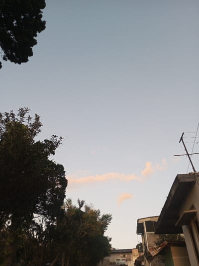 The image displays an outdoor scene in Sa Hti Law (3), Myanmar, during either dawn or dusk. The upper two-thirds of the frame are occupied by a pale blue sky, transitioning to a light, subtle pinkish-orange hue at a lower altitude, with scattered, elongated clouds exhibiting a similar faint tint. Darkly silhouetted tree canopies are prominent in the upper-left and lower-left sections, extending towards the center. On the right side, the upper portion of a building is visible, characterized by a dark, flat concrete roof overhang. An antenna structure, composed of a vertical pole with two horizontal cross-members, is affixed to this roof. Below the roof, a segment of a light-colored building wall with a dark rectangular outline, possibly a window or door frame, is discernible. Additional urban structures are faintly visible in the mid-background, partially obscured by the foreground vegetation. No individuals or discernible activities are present.