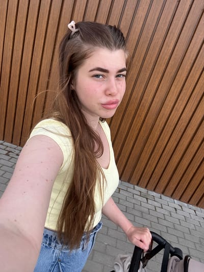 A young woman is taking a selfie while standing outside. She is wearing a yellow shirt and blue jeans. She has long brown hair. She is holding onto the handle of a stroller. The background includes a wooden wall and a brick walkway. The photo was taken in Речицы, Russia.