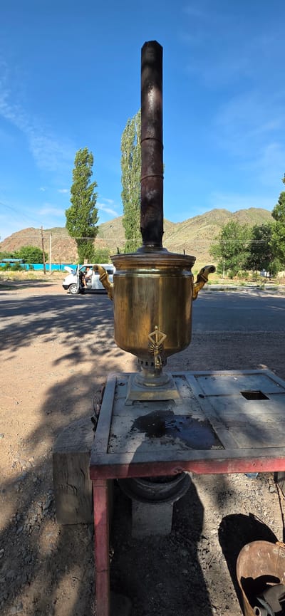 In Sasyk-Bulak, Kyrgyzstan, a brass samovar stands on a metal platform in the foreground. The samovar has two ornate handles and a spigot. A dark, cylindrical chimney extends upwards from the top of the samovar. The platform appears to be a makeshift structure with a red frame and a grey, perforated top. There are dark stains on the platform, suggesting recent use or liquid spillage.

Behind the samovar, a white van is parked on an asphalt road, with its hood open. A person is visible sitting in the driver's seat of the van. The ground beside the road is a mix of gravel and dirt. Tall, slender trees with green foliage stand in the middle ground. In the background, rolling hills with sparse vegetation and a clear blue sky with scattered wispy clouds complete the scene. The overall impression is an outdoor setting, possibly a roadside stop or a rural area.
