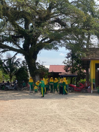 The image depicts a group of children, likely students, in a courtyard in Mekar Raya, Indonesia. They are dressed in green uniforms with yellow hijabs and sashes. The children are gathered around a large tree with moss-covered branches, and some appear to be interacting in a playful or instructional manner. To the right of the tree, a yellow building with a red-tiled roof is visible. Several motorcycles are parked on the gravelly ground, including a red one. The environment suggests an outdoor school setting or a public area with ample space. The sky is partly cloudy, indicating daylight.