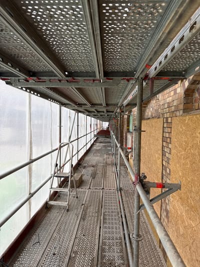 This image shows scaffolding on the exterior of a brick building. The scaffolding consists of metal platforms and railings. A ladder is also visible on the scaffolding. The building appears to be under construction or renovation, as some of the windows are covered with plywood. The location is not specified in Gothenburg, Sweden.