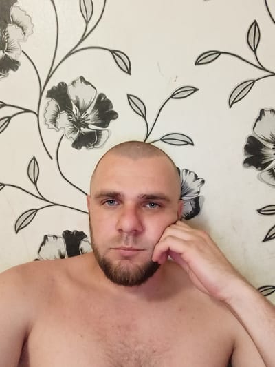 A shirtless man with a shaved head and a beard rests his head on his hand as he looks at the camera. He is positioned in front of a wall with floral wallpaper. The wallpaper features a repeating pattern of large, black and white flowers with thin black stems and leaves. The man's skin tone is fair, and his eyes are blue. His left arm is bent, with his hand supporting his cheek and jaw. The background appears to be an interior wall, possibly in a home. The image is a medium shot, focusing on the man's upper body and face.