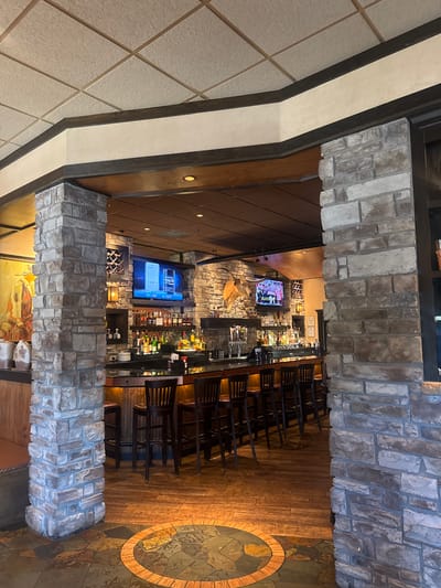 This image captures the interior of a restaurant or bar, taken from an elevated perspective looking down into the main bar area. The scene is framed by heavy stone pillars or arches, made of various shades of gray and brown rough-hewn stones, suggesting a rustic yet upscale ambiance.
The bar itself is well-lit and fully stocked with numerous bottles of liquor and spirits, arranged on shelves behind a dark counter. A series of dark wooden bar stools with slatted backs are neatly lined up along the bar. Above the bar, several flat-screen televisions are mounted. One screen prominently displays a mobile phone interface with text, showing "TEXT CODE TO: 213215" on the right side. Another screen shows what appears to be a sports event, with a figure in a uniform visible. On a wall near the bar, a mounted animal head, possibly an antelope or deer, contributes to the lodge-like decor.
The flooring in the bar area consists of dark wood planks, contrasting with a large circular decorative inlay made of different colored stone or tile, featuring a prominent brick-like ring. To the left, part of the ceiling is visible, made of textured, light-colored square tiles arranged diagonally. The overall setting appears to be a typical American casual dining or bar establishment, likely during off-peak hours as no patrons or staff are present.