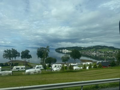 The image captures a view of a scenic waterfront campground in Hylla, Norway. Several white caravans are parked on a grassy slope overlooking a calm body of water. Trees are scattered across the landscape, with a cluster of larger trees on the left side of the frame. In the distance, a town with white buildings is visible along the opposite shore, nestled amongst rolling green hills. The sky is overcast with dramatic, textured clouds. A metal railing runs along the bottom of the foreground, suggesting the vantage point is from a road or elevated walkway. There are no people or animals visibly present in the scene. The overall atmosphere is serene and picturesque.
