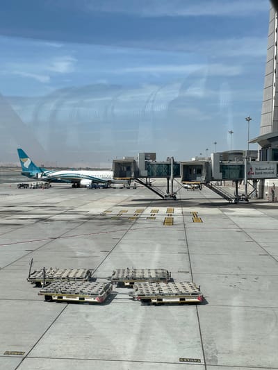 The scene depicts an airport apron at Muscat International Airport (MCT) in Seeb Province, Oman, under clear, bright daylight. An Oman Air passenger jet, identifiable by its white fuselage, teal and green tail fin with the airline's logo, and "OMAN AIR" livery, is parked at a gate. The aircraft is connected to two modern, grey and glass jet bridges, which extend from an adjacent terminal building. Each jet bridge includes external stairs descending to the tarmac.

Ground support equipment is present around the aircraft, including small service vehicles and what appear to be baggage carts. In the immediate foreground, four flat, rectangular cargo dollies with perforated surfaces are arranged on the concrete tarmac, which features various yellow and red operational markings. A section of a multi-story airport terminal building, displaying a "bank muscat" sign in both Arabic and English, is visible to the right. The sky is predominantly blue with scattered clouds. Reflections of interior architectural structures are evident on the glass surface through which the image was captured. Distant, light-colored, hazy hills are visible on the horizon. No distinct human figures are engaged in discernible activity.
