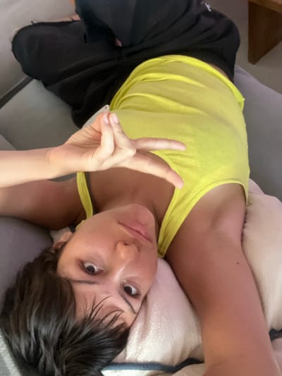 A woman with short dark hair lies on a pillow, wearing a bright yellow tank top and dark shorts. She is making a peace sign with her right hand, holding it up towards the camera. The image is taken from a slightly high angle, looking down at her face. Her eyes are open and looking directly at the camera. The background appears to be a sofa or bed.