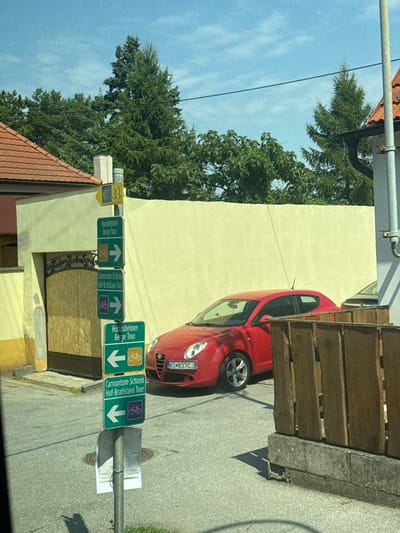 The image depicts a street scene in Wolfsthal, Austria, under a clear blue sky with scattered clouds. In the foreground, a red car, possibly an Alfa Romeo MiTo, is parked on the right side of the road, facing left. The car's license plate is visible as KS 837CJ. To the right of the car, a wooden fence is erected on a concrete base.

To the left of the car, a pole displays several green directional signs. The topmost sign, a yellow pilgrimage symbol, is affixed to the top of the pole. Below it are three green signs indicating bicycle tours. The first green sign, from top to bottom, reads "Hundsheimer Berge Tour" with a right-pointing arrow and a bicycle icon. The second sign reads "Carnuntum-Schloss Hof-Bratislava Tour" with a right-pointing arrow and a bicycle icon. The third sign reads "Hundsheimer Berge Tour" with a left-pointing arrow and a bicycle icon. A small, white paper is attached to the pole below the signs with blue tape.

Behind the car and the signs, a light yellow wall forms the backdrop. A large, ornate metal gate, partially covered with wooden planks, is visible to the left of the wall. Several trees are seen in the background, with a house with a red tile roof visible on the far left. The street itself is paved with asphalt. The scene appears to be a sunny day, likely during daylight hours.