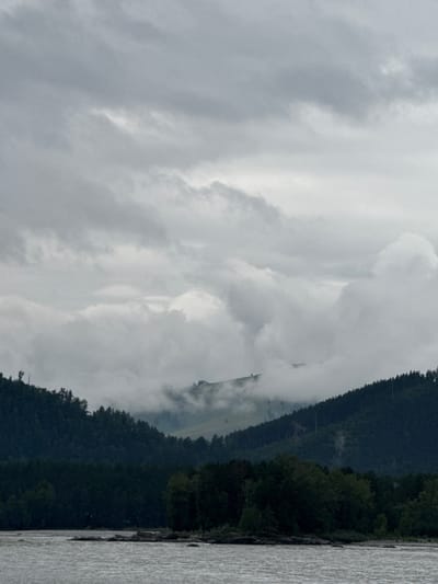 The image depicts a cloudy, overcast day over a river and forested hills. The foreground features a wide, choppy river with several dark rocks visible near the banks. Beyond the river, a dense, dark green forest covers the lower slopes of the hills. The hills in the middle ground are partially obscured by thick, white clouds that weave through the landscape, obscuring higher peaks. The sky is filled with heavy, grey clouds, with some lighter patches suggesting diffused sunlight. There are no people or man-made structures clearly visible in the frame. The scene conveys a sense of natural, rugged terrain under a somber sky. The location is identified as Nizhnekayancha, Russia.