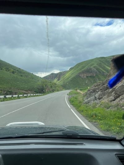 The image shows a view from inside a vehicle traveling on a winding road. The road is flanked by a steep, grassy mountain on one side and a rocky cliff with sparse vegetation on the other. A roadside barrier is visible along part of the road. The sky is overcast with clouds. The location is the city of Sisian, Armenia.
