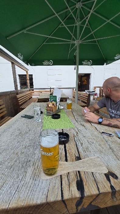The image shows a man sitting at a table on an outdoor patio of a restaurant in Micheldorf in Oberösterreich, Austria. He is wearing a gray t-shirt and is holding a pack of cigarettes. The man has short brown hair and a beard, and is wearing a watch on his left wrist. The table is made of wood and has a glass of beer on it. There are two other glasses of beer on the table, as well as a pack of cigarettes and a lighter. The table is under a large green umbrella, and there are wooden benches around the table. The patio is lined with a white wall and there is greenery in the background.  The image was taken at the coordinates latitude: 47.882, longitude: 14.13.
