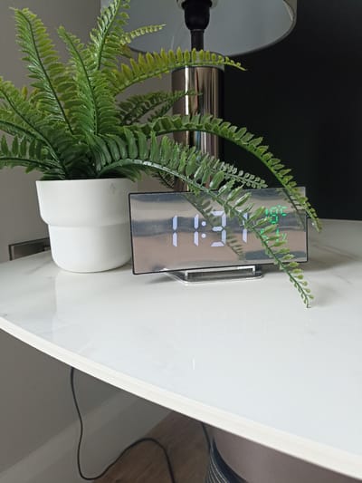 This is a medium shot, eye-level, indoor photograph of a white table with a white ceramic pot containing a fake fern plant, a chrome lamp base, and a digital alarm clock. The alarm clock displays the time 11:21 in white digital numbers, and in the upper right corner, it displays "10:05" in green digital numbers. The clock has a reflective, mirrored surface. The background is a dark wall, and to the left, a portion of a gray wall is visible. A black electrical cord snakes across the lower portion of the image. The photograph does not provide any information about the location in Doirí Beaga, Ireland.