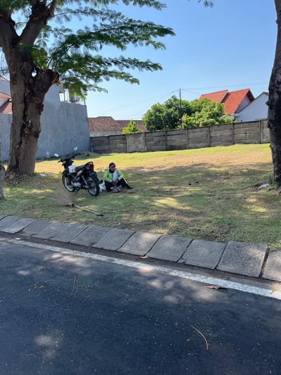In Entalsewu, Indonesia, a person is seen sitting on the grass near a motorcycle. The person, wearing a green long-sleeved shirt and a dark cap, is positioned to the right of a parked motorcycle. A broom lies on the ground in front of the motorcycle. The scene is set in an outdoor area with a concrete wall and houses in the background, under a clear blue sky. The ground is covered in grass, with trees visible on the left and right sides of the frame. A road with a concrete curb is in the foreground.