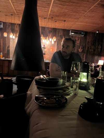 A man sits at a table set with glassware, dishes, and a lantern. The table is draped with a light-colored cloth. The location is in Livingstone, Zambia. The background shows interior walls with wooden and woven textures, illuminated by multiple hanging lights.