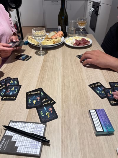 The image depicts a table setting with a card game in progress. On the table are cards from a game called "Wizard," a scorekeeping pad, a pen, glasses of wine and water, and plates of food including bread rolls and charcuterie. Two individuals are visible, one holding a hand of cards and the other's hand is visible resting on the table. A cigarette pack is also present on the table. The setting appears to be indoors, likely a dining area within a residence. The specific location of Langeoog, Germany, is not identifiable from the visual information provided in the image.