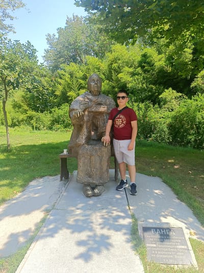 A young boy stands to the right of a bronze statue of a seated, elderly woman. The boy is wearing a maroon t-shirt with a circular logo, light-colored shorts, and black and white sneakers. He has dark hair and is wearing sunglasses. The statue depicts the woman with her hands resting on her lap. A small wooden bench is positioned behind the statue. In front of the statue is a stone plaque with Bulgarian text. The scene is outdoors, with lush green trees and grass surrounding the monument. Sunlight filters through the leaves, casting dappled shadows on the ground, indicating it is daytime, likely a sunny day. The overall setting appears to be a park or memorial site. The text on the plaque translates to: "VANGA. The sculpture was made on the occasion of the 100th anniversary of the birth of the Prophetess, by the sculptor Emil Popov, and is a donation from him to the 'Vanga' Foundation, 3.X.2011."