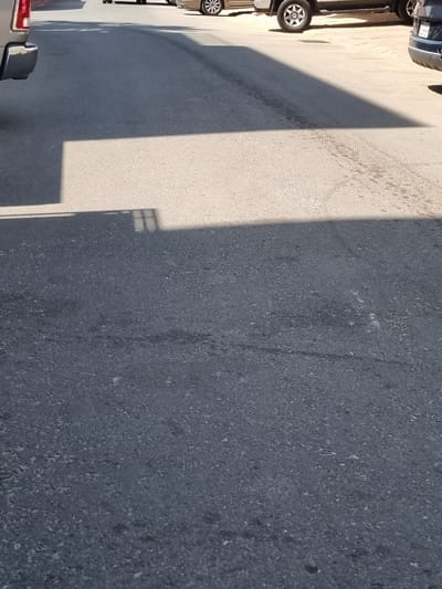 A low-angle, vertical outdoor photograph shows a paved asphalt road surface in the city of Rumaithiya, Kuwait. A shadow of a building, with a grid-like pattern, is cast across the road. The road is dark gray and shows signs of wear and tear, with some lighter patches and tire marks. In the upper portion of the image, the wheels of parked vehicles are visible, suggesting a parking lot or street. The lighting indicates it is daytime, likely sunny, given the sharp shadow.