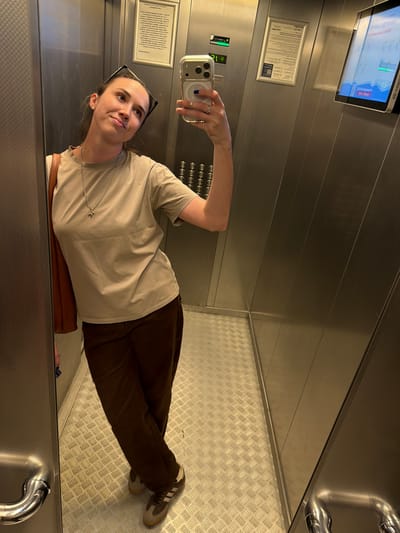 A young woman is taking a selfie in an elevator. She is wearing a light beige t-shirt, brown pants, and grey sneakers. She has on a silver necklace with a small cross pendant and a pair of black-framed glasses perched on her head. The elevator walls are made of brushed metal, and the floor is a diamond-patterned metal. There is a display screen on the upper left wall showing some information, and a framed document with text below it. The woman appears to be looking up and slightly to the left, with a slight smile on her face, suggesting a candid or playful moment. The overall lighting is artificial and diffused, typical of an indoor elevator. The setting suggests a modern building or public space in Elizavetinskaya, Russia.