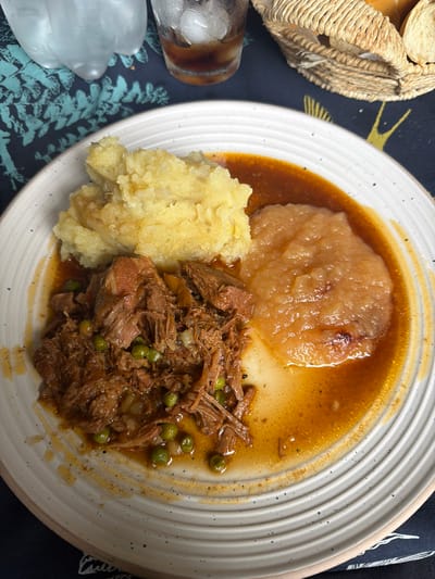 A top-down view shows a white plate containing three separate mounds of food. At the top of the plate is a large scoop of applesauce, surrounded by a light brown gravy. To the left is a pile of mashed potatoes. To the right of the potatoes is a pile of shredded meat with some green peas mixed in. Some of the meat and peas are sitting in the gravy. To the upper left of the plate is a bread basket filled with bread slices. To the left of the plate and below the bread basket is a glass with a brown liquid and ice. The plate is on a dark blue tablecloth with a light blue fern-like pattern. Parts of a black cloth are visible in the upper right. There is no indication this photo was taken in La Lucila, Argentina.