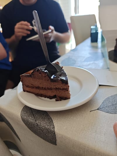 A slice of chocolate cake sits on a white plate on a tablecloth with a leaf pattern. A spoon is stuck in the cake. A person in a dark t-shirt is using a phone or camera in the background. Another person in a blue shirt is partially visible on the left. The setting appears to be indoors, possibly a dining room. There is a blurred chair and box in the background. The location cannot be determined beyond the general area of Heraklion Municipal Unit, Greece.