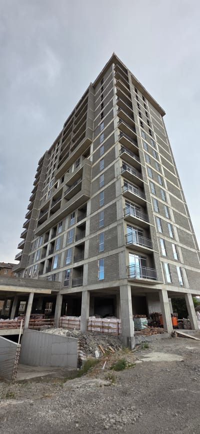 An unfinished, multi-story building is shown from a low-angle perspective against a cloudy sky. The building is constructed primarily of concrete blocks with concrete pillars and horizontal concrete bands. It has numerous balconies with black metal railings and rectangular windows, some of which are already glazed. Construction materials, including pallets of bagged materials and wooden planks, are visible at the base of the building. The ground in the foreground is a mixture of gravel and dirt, with some vegetation. In the background, another, older building with windows and a pitched roof can be seen. The overall scene depicts an ongoing construction project in the city of Gori, Georgia.