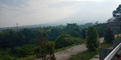 The image displays an elevated, hazy landscape in Cugenang, Indonesia. In the foreground, a grey guardrail is partially visible on the right, bordering a curving, unpaved path that recedes into the midground. Two small, slender trees, one with green leaves and the other with reddish foliage, stand to the left of this path. The midground is dominated by a dense, varied canopy of dark green trees. On the far right, a two-story grey building with a reddish-brown tiled roof is partially visible, nestled within the foliage. The background features a large mountain peak, indistinct due to atmospheric haze, dominating the left side of the frame. Several metal power transmission towers are discernible above the distant tree line, extending towards the right. The sky is uniformly light grey, indicative of overcast conditions or significant atmospheric haze. No individuals or active subjects are present in the scene.