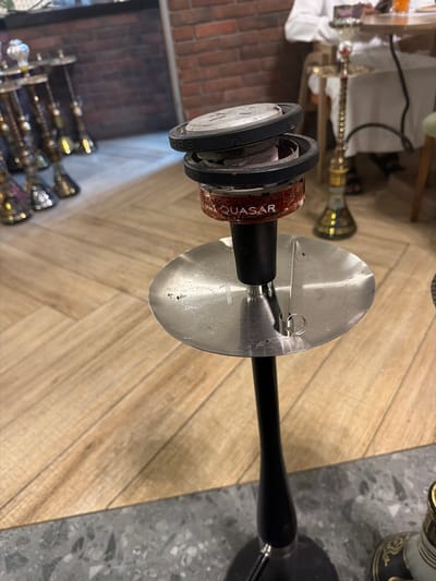 This image captures an indoor scene within a lounge or cafe located in Business Bay, United Arab Emirates. The primary subject is a modern hookah, positioned centrally in the foreground. It features a black, tapered stem and a circular, brushed stainless steel ash tray with a small, metallic pair of tongs hanging from its edge. Above the ash tray, a transparent red glass bowl, labeled "QUASAR" in white text, contains visible red tobacco. Resting on this bowl is a black heat management device, open to reveal two pieces of glowing charcoal.

The floor transitions from a light-toned wooden plank surface, visible in the mid-ground and extending to the left, to a darker, grey terrazzo-patterned tile in the immediate foreground, located beneath the hookah's base.

In the left background, several traditional hookahs with ornate metallic designs and clear bases are arranged, out of focus, on the wooden floor. A red brick wall is partially visible behind them. In the right background, another traditional hookah stands near a wooden table where an individual, wearing a white thobe, is seated. An orange glass is visible on the table adjacent to the person.