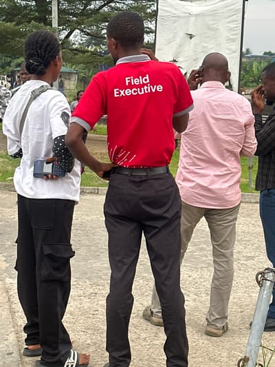 Three individuals stand closely together. The person in the foreground wears a white shirt, black pants with side pockets, and sandals. They hold a calculator-like device. Behind them, a person in a red shirt labeled "Field Executive" wears black pants and a belt. The third person wears a pink striped shirt and tan pants. They appear to be engaged in a phone call. A white tarp or banner is visible in the background, partially obscuring some trees. The setting appears to be outdoors, possibly a parking lot or paved area.