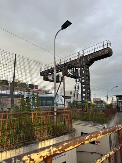 The image shows a section of railway infrastructure in Saint-Brice-sous-Forêt, France. A large, dark, steel gantry, possibly used for signaling or electrification, dominates the frame. It is constructed of riveted metal, with cylindrical openings cut into its vertical support. A metal ladder ascends the structure. A modern streetlight with a rectangular LED fixture stands nearby.
In the foreground is a rusty, dilapidated metal fence overgrown with vegetation. A chain-link fence extends further back, obscuring the view.
Beyond the fences are railway tracks and an SNCF passenger train visible through the fencing. The sky is overcast and gray.
